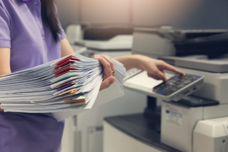 Bussinesswoman using copier machine to copy heap of paperwork in office.